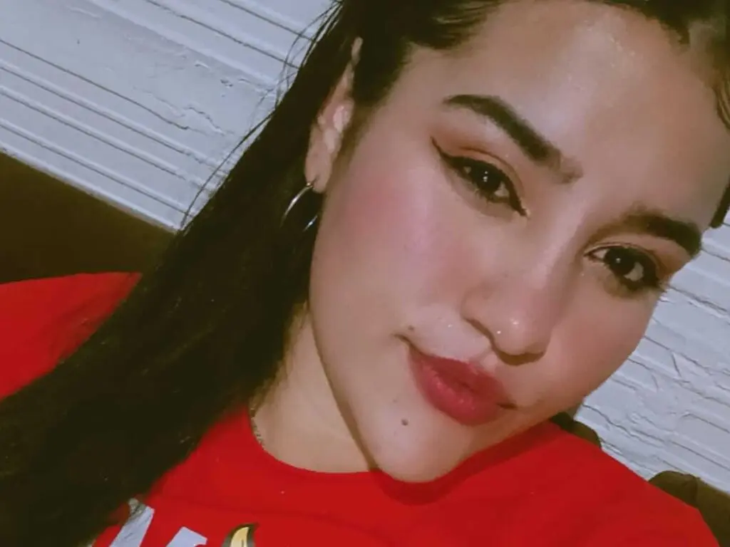 Young woman with winged eyeliner and red t-shirt posing for a close-up selfie in a dimly lit room.