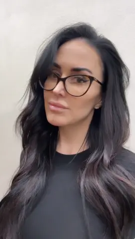 Brunette woman with long dark hair and glasses wearing a black top against a neutral wall background.