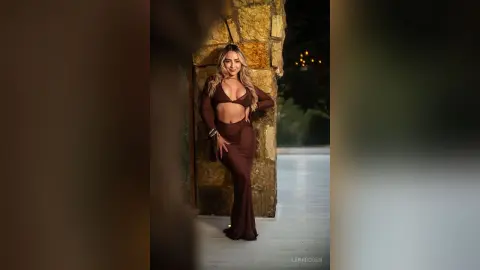 Glamorous woman in a chocolate brown two-piece outfit posing against a rustic stone wall in a moody setting.