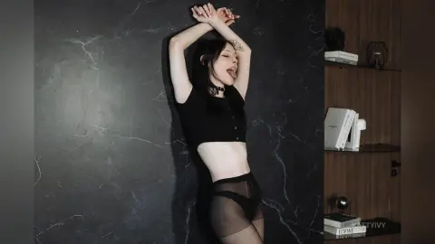 Young woman in black crop top and sheer tights posing playfully against a dark marble wall in a moody setting.