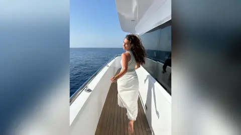Woman in a white summer dress standing on a luxury yacht deck overlooking the blue ocean under a clear sky.
