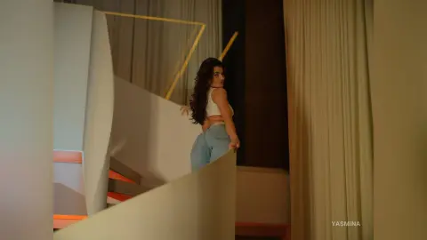 Young woman in a white crop top and denim jeans posing on a modern staircase in warm indoor lighting.