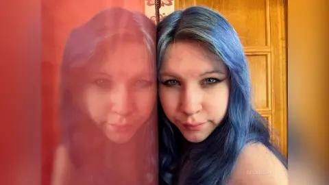 DeidreSarin poses with her long blue hair against a wooden door, looking directly at the camera with a soft expression.