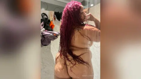 Curvy woman with long wavy red hair posing nude in a bright modern bathroom setting.