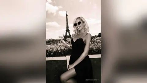 NickyAshcroft smiles in a black slip dress and sunglasses, holding a glass of wine with the Eiffel Tower in the background.