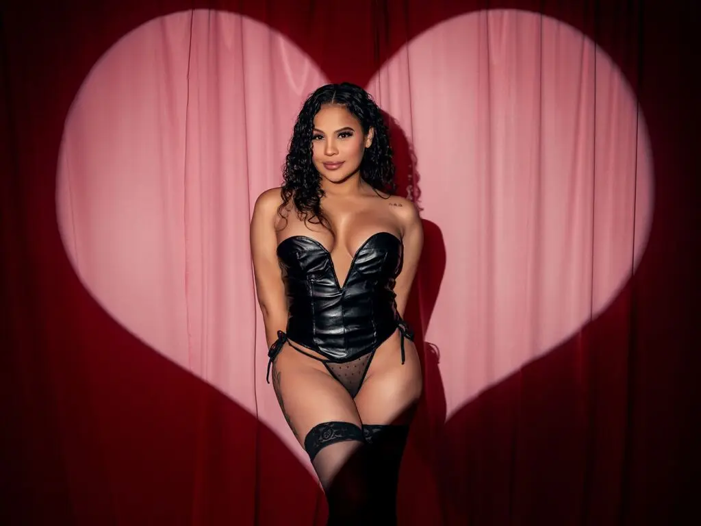 Curvy woman in black leather corset and lace stockings posing against a romantic pink heart backdrop.