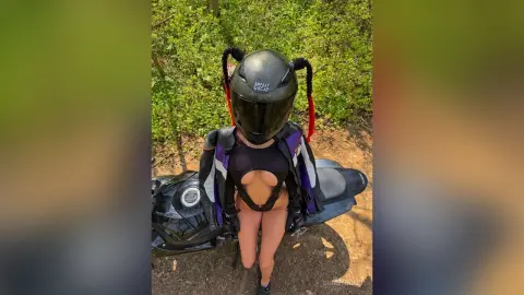 Woman in black bodysuit and motorcycle gear sitting on a black bike in a sunny forest setting.