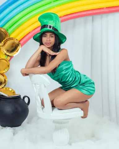 Smiling woman in a green satin dress and leprechaun hat sitting by a rainbow and pot of gold.