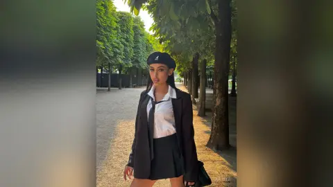 Stylish woman in a black blazer, white shirt, and beret standing on a sunny tree-lined path.