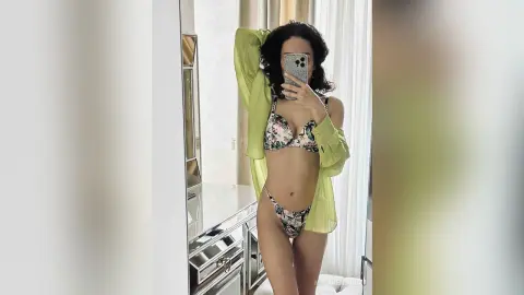 Young woman in floral bikini and lime green shirt posing for a mirror selfie in a bright modern room.