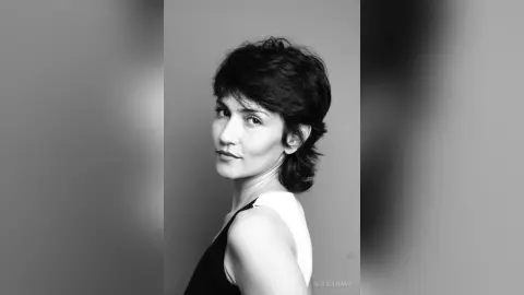 IveteDurand looks over her shoulder with a direct gaze, showing her short dark hair and black tank top against a plain background.