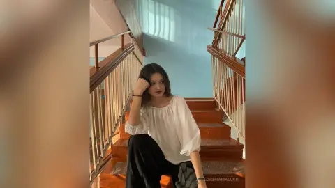 Young woman in a white blouse sitting on wooden stairs in a bright, minimalist interior setting.