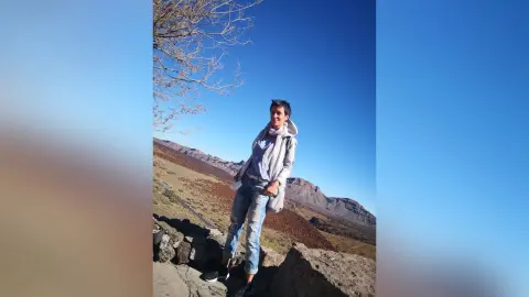 Smiling young woman in a grey hoodie and jeans standing on a rocky mountain trail under a clear blue sky.