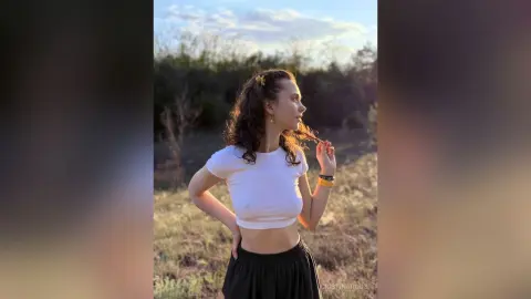Young woman in a white t-shirt and black skirt posing in a sunny outdoor field at sunset.