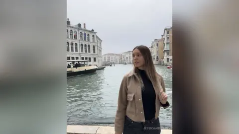 JessicaAidennn — Young woman in a tan jacket and black top standing by a scenic Venice canal on an overcast day.
