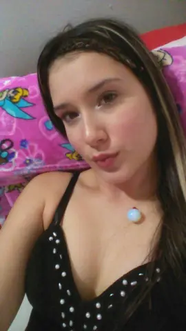 Young woman with long brown hair wearing a black studded tank top relaxing in a bedroom setting.