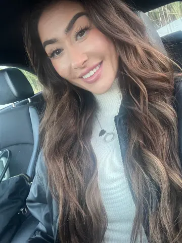 OliviaEdenBabestation smiles while sitting in a car, wearing a white turtleneck and a black jacket with her long brown hair down.