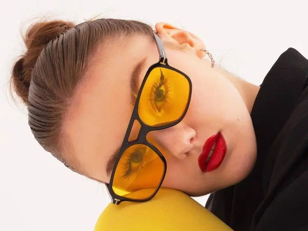 Fashionable woman with red lips and yellow tinted sunglasses posing in a black outfit against a white background.