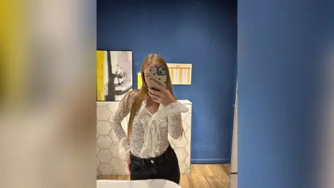 AdenaMcfalls poses in a mirror wearing a white lace long-sleeve blouse and black jeans, showing her long blonde hair.
