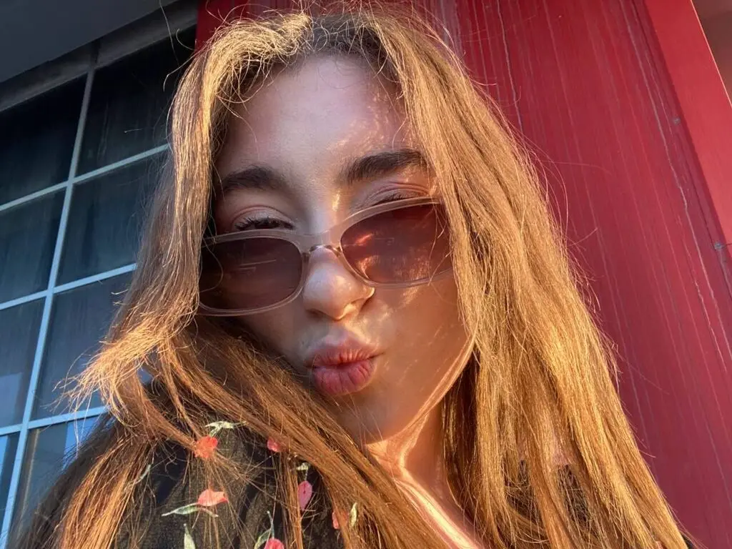 Young woman with long hair wearing tinted sunglasses and a floral top in warm golden hour sunlight.
