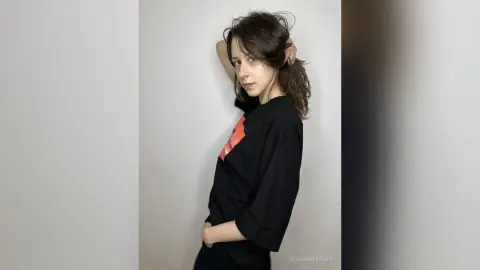 Young woman with wavy brown hair wearing a black graphic t-shirt posing against a white wall in a minimalist style.