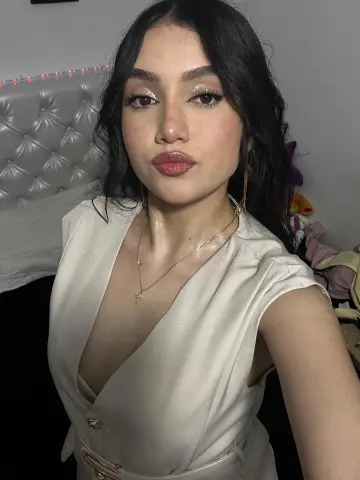 Young woman with glamorous makeup wearing a white v-neck top and gold necklace in a cozy bedroom setting.
