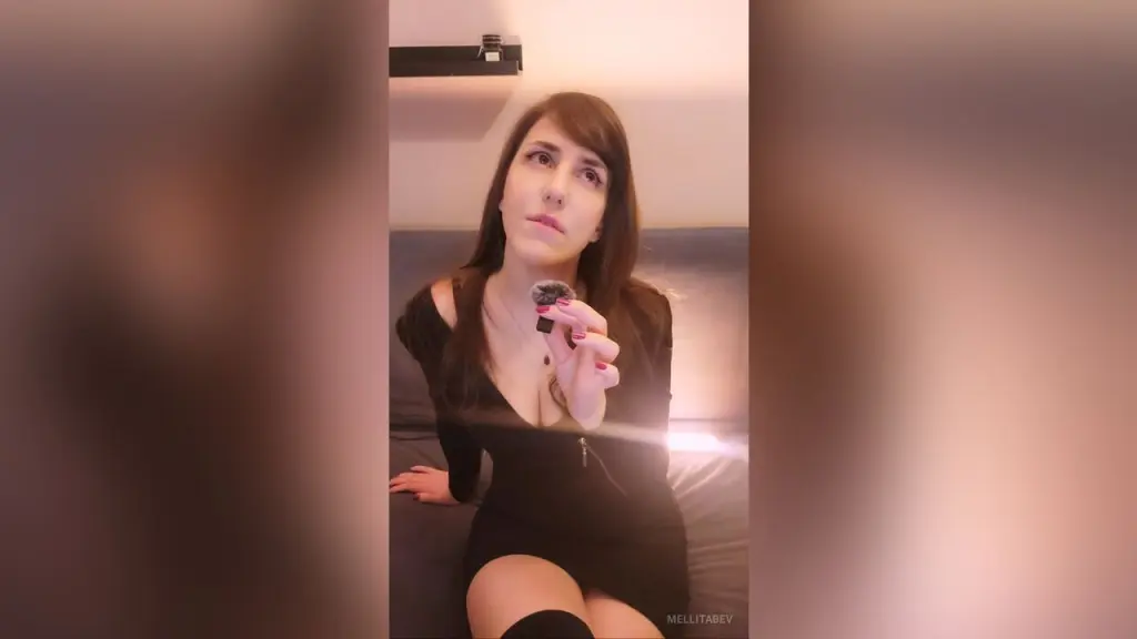 Young woman in a striped tank top and black skirt kneeling on a white bed in a bright, cozy bedroom.