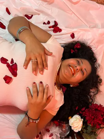 GenesisMontenegro laughs while lying on white sheets covered in red rose petals, wearing a white top and jewelry.