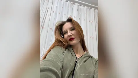 Redheaded woman with glasses in a green hoodie posing indoors against white curtains with a relaxed mood.
