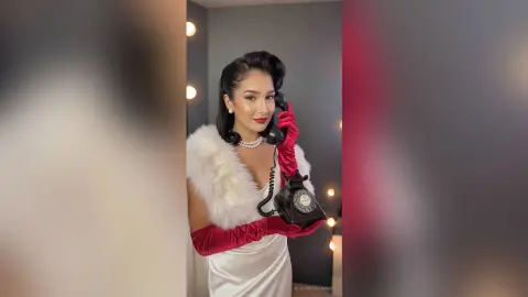 Glamorous woman in a white satin dress and red gloves holding a vintage phone in a retro studio setting.