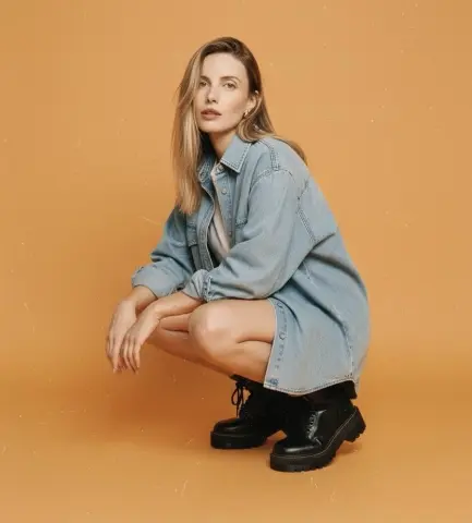 Young woman in a light blue denim shirt and black boots posing against a warm tan background.