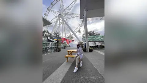 Stylish woman in a white blazer and beige trousers sitting at a wooden picnic table near a giant Ferris wheel.
