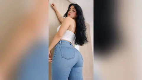 Young woman in white tank top and blue denim jeans posing against a neutral wall with a confident mood.