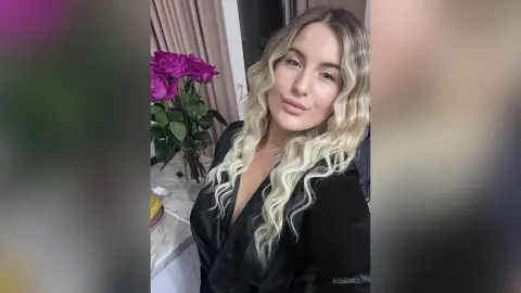 Blonde woman in a black satin robe smiling in a bright room next to a bouquet of pink roses.