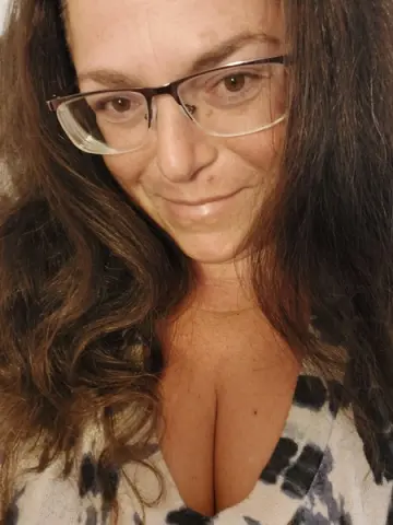 Smiling woman with long wavy hair and glasses wearing a white patterned top in a warm indoor setting.