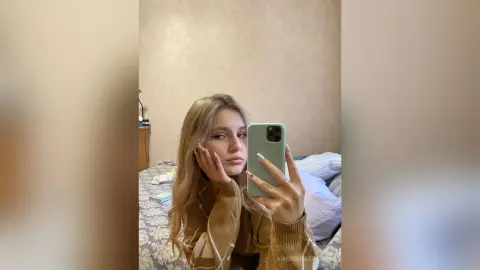 Young woman with long blonde hair taking a cozy selfie in a beige sweater at home.
