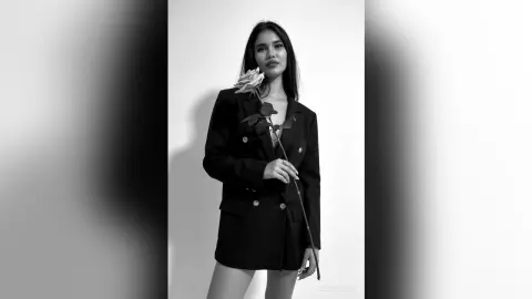 Elegant woman in a black blazer holding a single rose in a moody black and white studio portrait.