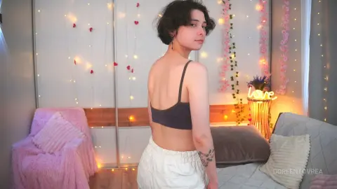 Young woman in a grey crop top and white shorts posing in a cozy bedroom with pink fairy lights and romantic decor.
