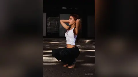 Stylish woman in a white tank top and black trousers posing in a moody, sunlit urban concrete setting.