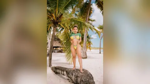 Slim woman in a green bikini posing on a tropical beach under palm trees during a sunny summer vacation.