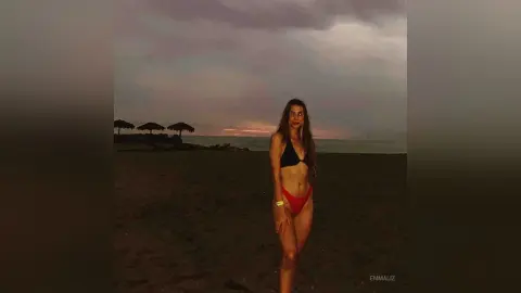 Young woman in a black bikini and red bottoms standing on a dark beach under a moody twilight sky.