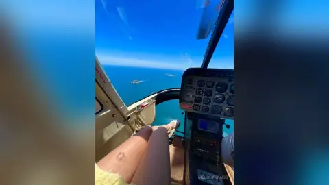 View from a helicopter cockpit overlooking a bright blue ocean and coastline on a sunny day.