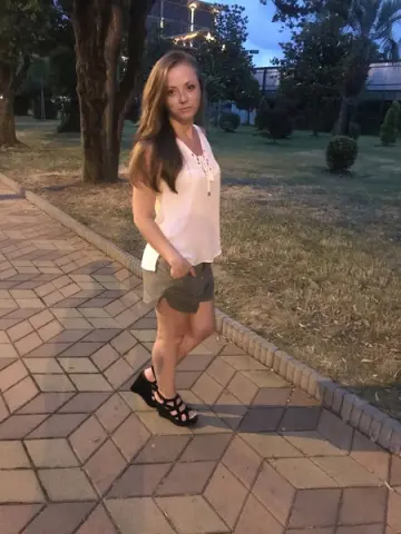 Young woman in a white sleeveless top and olive shorts standing on a stone path in a park at sunset.