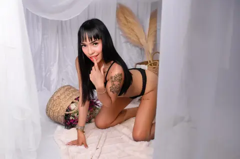 Smiling woman with black bangs in black lingerie posing on a white rug in a dreamy, bright bohemian bedroom.