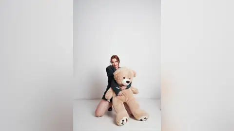 AryaSin smiles while hugging a large, tan teddy bear, wearing a black long-sleeved top and sitting on a white floor.