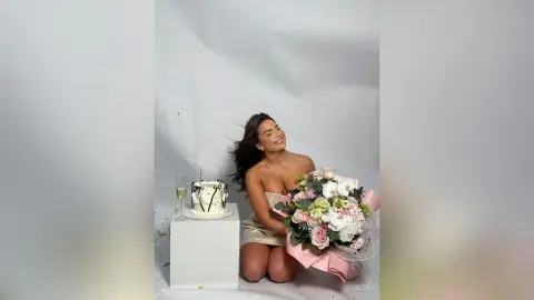 Smiling woman in a beige dress holding a large bouquet of pink roses next to a cake in a bright studio setting.