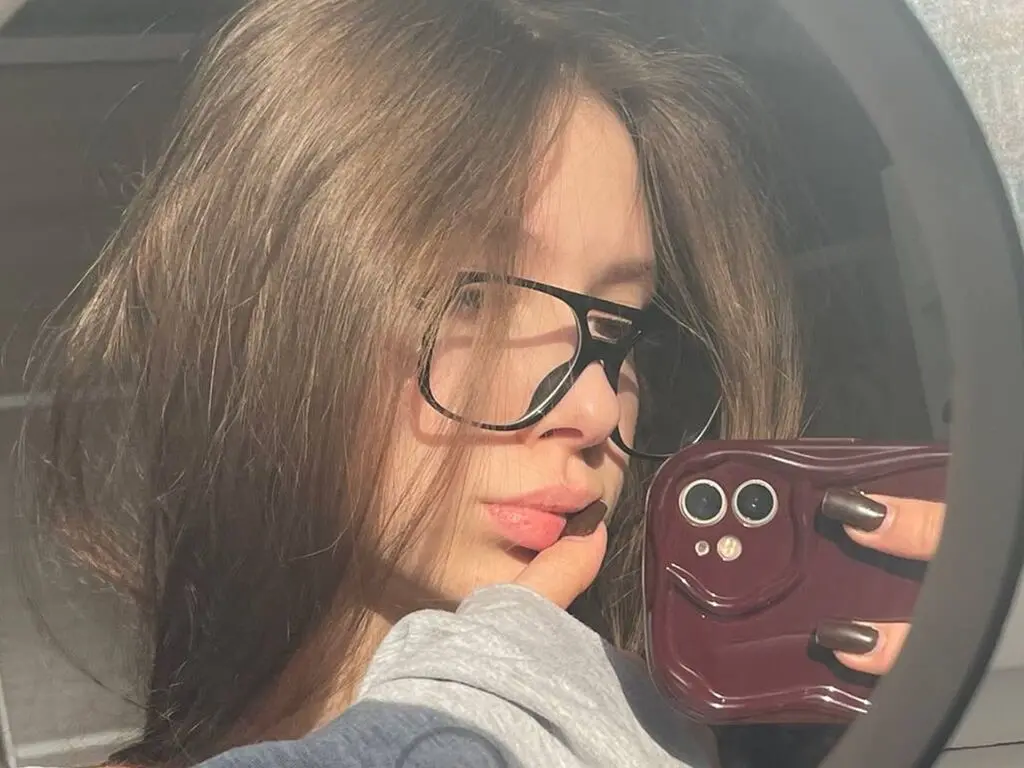 Young woman with long brown hair wearing black glasses taking a casual mirror selfie in soft sunlight.