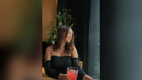Smiling young woman in a black off-the-shoulder top relaxing by a window with indoor plants in a cozy setting.