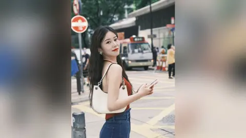 Young woman in a brown tank top and jeans holding a phone on a sunny city street.