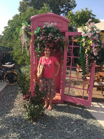 Smiling woman in a pink shirt and floral skirt standing by a pink flower-covered phone booth in a sunny garden.